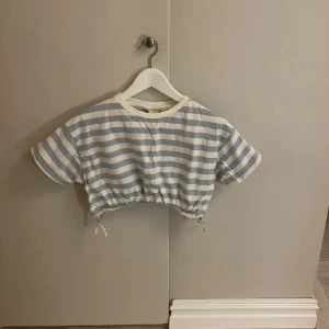 Randig croppad topp från Zara - Superfin croppad topp från Zara med blå och vita ränder. T-shirten har korta ärmar, rund halsringning och dragsko med knyt i midjan för en snygg passform. Perfekt för dig som gillar en chill och trendig stil.