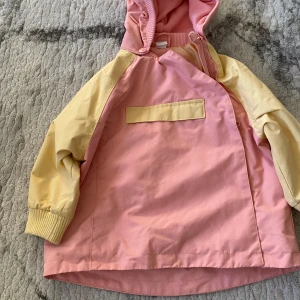 Pastellrosa och gul vindjacka med huva - Söt vindjacka i pastellrosa med ljusgula ärmar och detaljer. Jackan har luva, dragkedja och en stor ficka framtill med gul flik. Perfekt för blåsiga dagar och lek utomhus. 