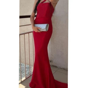 Röd långklänning med smala axelband - Supersnygg röd långklänning med smala axelband och figurnära passform. Klänningen har markerad midja, lätt glansigt tyg och en elegant, släpande nederdel. Perfekt för bal eller festkvällar när du vill sticka ut.