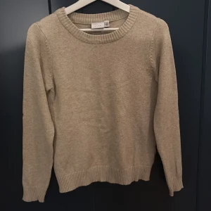 Beige stickad tröja från Vila - En klassisk beige stickad tröja från Vila med rund halsringning och ribbade muddar vid ärmslut och nederkant. Perfekt för lager-på-lager och enkel att matcha med jeans eller kjol. Långärmad och i mjukt material som känns skönt mot huden.
