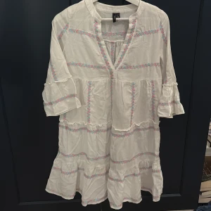 Vit  kortklänning med volanger - Superfin vit kortklänning från Vero Moda med broderade pastellfärgade detaljer i rosa och blått. Klänningen har volanger, trekvartsärm och v-ringning. Perfekt för sommarens alla tillfällen och ger en avslappnad boho-vibe.