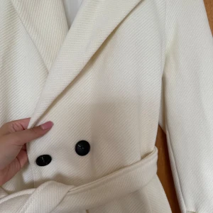 Vit kappa med bälte och knappar - Stilren vit kappa med bred krage, svarta knappar och knytskärp i midjan. Lång modell med klassisk look och snygga detaljer på ärmsluten. Perfekt för dig som vill ha en clean och elegant vibe. Använd en gång.