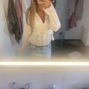 Vit blus med volang och v-ringning - Supersöt vit blus med v-ringning och volangdetaljer. Lätt struktur i tyget och långa ärmar ger en romantisk vibe. Perfekt till jeans eller kjol för en fräsch look. 