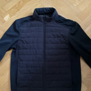 Mörkblå carigan från ZARA - Snygg svart quiltad jacka från Zara med dragkedja framtill. Jackan har en stilren design med ståkrage och långärmade ärmar. Perfekt för dig som gillar en clean look och vill hålla värmen under kyliga dagar.