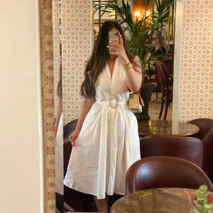 Vit omlottklänning med bälte - Säljer en vit, ärmlös klänning med v-ringning och markerad midja med brett bälte och rund spänne. Klänningen har knappar framtill och ett luftigt, lätt utställt kjolparti som slutar strax under knäet. Perfekt för varma dagar och stilrena outfits.