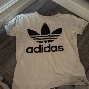 Vit Adidas t-shirt med stor logga - Vit t-shirt från Adidas med den klassiska stora svarta trefoil-loggan och adidas-tryck på bröstet. T-shirten är kortärmad och gjord i mjuk bomull, perfekt för en avslappnad streetstyle-look.