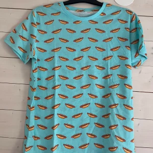 Turkos t-shirt från biltema med hotdog-mönster - Turkos t-shirt från biltema med ett heltryckt, lekfullt hotdog-mönster över hela plagget. Klassisk rund hals och korta ärmar. Perfekt för dig som gillar färg och vill sticka ut. Skön och mjuk känsla, troligtvis i bomull.