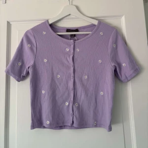 Lila blommig topp från Primark - Supergullig lila topp från Primark med små broderade blommor över hela. Kortärmad, croppad modell med rund hals och knappar framtill. Perfekt för dig som gillar söta detaljer och en chill vibe. Aldrig använd, skulle mer säga att den är strl S.