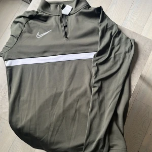 Nike Dri-FIT grön half zip tröja - Snygg grön långärmad tröja från Nike med vit logga och vit rand över bröstet. Tröjan har half zip och är gjord i Dri-FIT polyester, perfekt för träning eller chill. Skön passform och sportig look, funkar året runt. Skick 10/10.