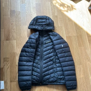 Svart dunjacka Peak Performance S - Säljer en svart dunjacka från Peak Performance i storlek S. Jackan har huva, dragkedja och klassisk quiltad design. Perfekt för kyliga dagar och riktigt skön att bära. Diskret logga på bröstet och ärmen. Snygg och stilren look.