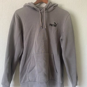 Grå hoodie från Puma med huva - Säljer en stilren grå hoodie från Puma med klassisk logga på bröstet. Tröjan har huva med snörning och en stor magficka framtill. Perfekt för chill dagar eller när du vill ha en enkel och snygg look.