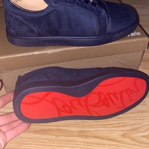 Mörkblå Louboutin sneakers mocka - Exklusiva mörkblå sneakers från Louboutin i lyxig mocka med signaturröd sula och broderad logga på hälen. Snygga rundade tår, platt sula och klassisk snörning. Perfekta för dig som vill sticka ut med stilrena detaljer och premiumkänsla.