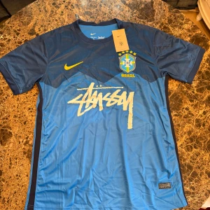 Stüssy Nike fotbollströja - Exklusiv Brasilien fotbollströja från Nike i samarbete med Stüssy. Tröjan är blå med mörkare blå detaljer, har korta ärmar och är tillverkad i Dri-FIT material. Stort Stüssy-tryck framtill, Brasil-logga och Nike Swoosh i gult.