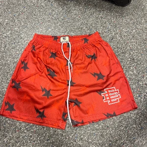 Röda meshshorts från Eric Emanuel - Snygga röda meshshorts från Eric Emanuel med svarta stjärnmönster och vit logga på benet. Shortsen har elastisk midja med vit snörning och är tillverkade i luftig polyester, perfekta för sommaren eller träning.