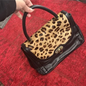 Svart handväska med leopardmönster - Cool svart handväska i skinn med lock i leopardmönstrad mocka. Väskan har ett rejält handtag, snyggt spänne i metall och flera fack inuti för bra organisering. Perfekt för dig som vill sticka ut med en trendig accessoar.