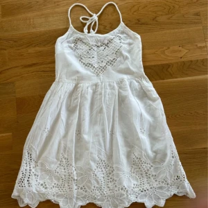 Vit somrig klänning med spetsdetaljer - Superfin vit kortklänning med tunna axelband och knyt i nacken. Klänningen har broderade blommor och spetsdetaljer både upptill och längs fållen. Perfekt för varma dagar och har en luftig känsla tack vare bomullsmaterialet.