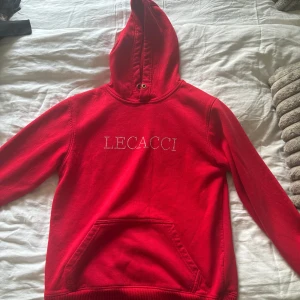 Röd hoodie med strassfjäril från Lecacci - Röd hoodie från Lecacci med snygg strasslogga framtill och en stor fjäril i strass på ryggen. Tröjan har huva, magficka och långa ärmar. Perfekt för dig som gillar statement-plagg och vill sticka ut lite extra.