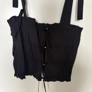 Svart korsettopp med snörning Zara XS - Supersnygg svart korsettopp från Zara i storlek XS. Toppen har breda axelband, smockad struktur och snörning framtill med små glittriga detaljer. Volangkant nertill och en söt rosett på ena axelbandet. Perfekt för dig som gillar trendiga detaljer och en edgy look.