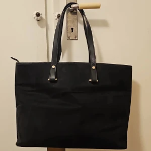 Svart axelväska med skinnhandtag - Stilren svart axelväska med rejäla handtag i skinn och diskreta guldfärgade detaljer. Väskan har en klassisk, fyrkantig form och gott om plats för allt du behöver till skolan eller stan. Perfekt för dig som gillar enkel och tidlös design. Om man vill ha fler bilder är det bara att skriva. 