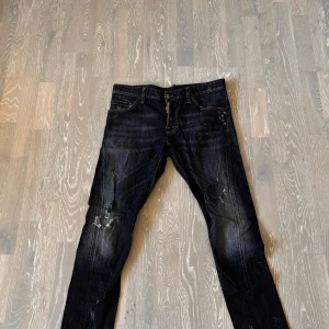 Mörkblå Dsquared2 jeans med slitningar - Snygga mörkblå jeans från Dsquared2 med slitna detaljer och  färgstänk. Klassisk femficksmodell med läderpatch bak i midjan. Jeansen har en smal passform och är tillverkade i stretchig denim för extra komfort.