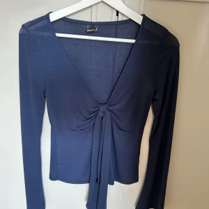 Mörkblå omlottopp från Gina Tricot - Supersnygg mörkblå omlottopp från Gina Tricot med lång ärm och djup v-ringning. Toppen har knyt framtill som ger en trendig look och är gjord i ett mjukt, stretchigt material. Perfekt att styla med jeans eller kjol för en cool vibe.