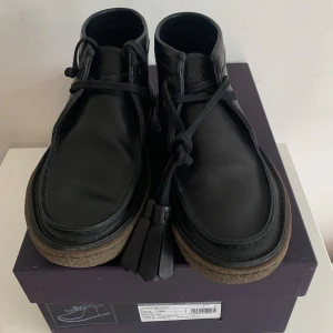 Svarta kängor från Prada i skinn - Stilrena svarta kängor från Prada i äkta skinn med grov crepesula och snörning med dekorativa tofsar. Klassisk rund tå och diskret logga på plösen. Perfekta för dig som vill ha en clean och lyxig look med edge.