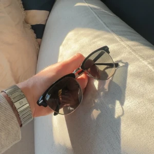 Svarta Ray-Ban Clubmaster glasögon - Snygga Ray-Ban Clubmaster med svart ovandel och silvriga detaljer. Klassisk halvbåge och retrovibbar, perfekt för dig som vill sticka ut. Tidlös design med genomskinliga glas och metallbåge undertill.