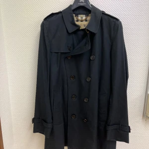 Burberry Men’s Heritage Kensington Short Trench Coat  - Storlek: 50 EU (motsvarar Medium–Large) 	•	Färg: Marinblå 	•	Modell: Kensington – kort längd 	•	Tillverkad i England 	•	Dubbelknäppning, axelklaffar, stormflik, justerbara ärmslut, bälte med metallspännen (D-ringar) och klassiskt rutigt Burberry-foder 	•	Skick: Mycket fint begagnat skick, sparsamt använd och väl omhändertagen 	•	Alla originaldetaljer finns kvar  Nypris ca 23 500 kr