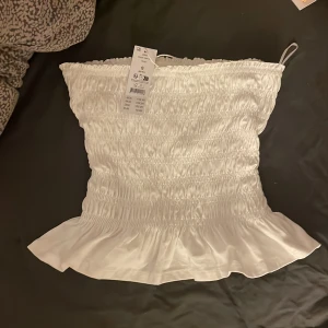 Vit tubtopp med volang från Gina Tricot - Supersöt vit tubtopp från Gina Tricot med smockad design och volangkant nedtill. Toppen är axelbandslös och har en stretchig passform som sitter snyggt. Perfekt för att skapa en fräsch och trendig sommarlook den är helt ny aldrig aldrig använd!❤️