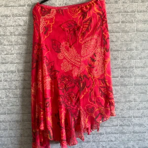 Röd mönstrad långkjol från Steilmann - Färgstark långkjol från Steilmann i rött med blommigt och paisleymönster i orange och svart. Kjolen har asymmetrisk volangkant och detaljer med pärlor längs vissa blad. Tunt, flowy tyg som ger en riktigt snygg siluett. Storlek 44 står det men jag skulle säga att den passar 36-40. 