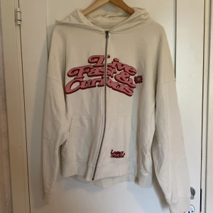 Vit hoodie från Legacy Studios - Säljer en vit hoodie från Legacy Studios med dragkedja och huva. Framsidan har ett stort tryck i rosa och rött med texten 'Live Fast & Curious' och en mindre logga vid fickan. Hoodien är i mjuk bomull och har långa ärmar och känguruficka. Pris går attt diskuteras!