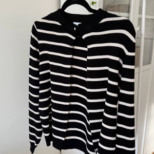 Randig svartvit stickad tröja - Snygg svart och vit randig stickad tröja med lång ärm och rund halsringning. Tröjan har en cool öppning framtill med knappdetalj vid halsen. Perfekt för dig som gillar klassiska mönster med en twist.