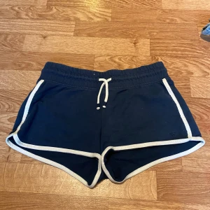 Marinblå shorts från Hampton Republic - Snygga marinblå shorts från Hampton Republic med vita kanter och snörning i midjan. De har en sportig look med elastisk midja och kort passform, perfekta för varma dagar. Materialet är mjukt och skönt, troligtvis bomull. Passar dig som gillar en avslappnad och trendig stil.