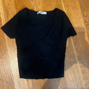 Svart omlottopp från Zara  - Svart ribbad omlottopp från Zara  med korta ärmar och djup v-ringning. Snygg croppad modell med markerade sömmar som ger en cool look. Perfekt att styla med höga jeans eller kjol.