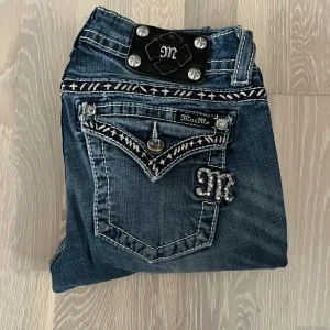 Miss Me jeans med broderade detaljer - Snygga blå Miss Me jeans med vita broderade detaljer och dekorativa knappar på fickorna. Jeansen har tydliga kontrastsömmar och coola detaljer på bakfickorna. Klassisk femficksmodell i jeansmaterial. Ena knappen på en av bakfickorna är utbytt