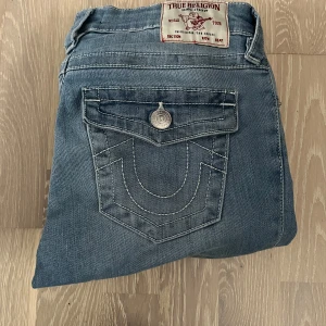 True Religion Becca bootcut jeans - Snygga ljusblå jeans från True Religion, modell Becca med bootcut och midwaist. Klassiska kontrastsömmar, ikonisk fickdesign och silverfärgade knappar. Tillverkade i mjukt denim för en skön känsla och cool look.