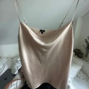 Champagnefärgat linne - Säljer en stilren champagnefärgad linnetopp från River Island med tunna justerbara axelband och snygg draperad halsringning. Toppen har en slät satinliknande yta som ger en lyxig känsla och passar perfekt till jeans eller kjol.