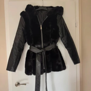 Svart pälsjacka med bälte och huva - Snygg svart pälsjacka med skinnärmar och huva. Jackan har dragkedja framtill och ett matchande bälte i midjan för extra stilpoäng. Perfekt för dig som vill ha en statementjacka med både päls- och skinnkänsla. Fuskpäls jacka 