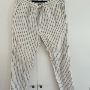 Randiga linnebyxor från H&M - Snygga vita linnebyxor från H&M med svarta vertikala ränder och dragsko i midjan. Regular fit med elastisk midja och fickor både fram och bak. Perfekta för en chill och stilren look under varmare dagar.