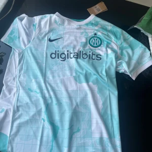 Inter Milan bortatröja 2022/23 Nike - Säljer en Inter Milan bortatröja från säsongen 2022/23. Tröjan är vit med ljusblå kartmönster och har klubbmärket och Nike-loggan på bröstet. Digitalbits som sponsor på framsidan. Tillverkad i lätt och ventilerande material, perfekt för fotboll eller streetwear.