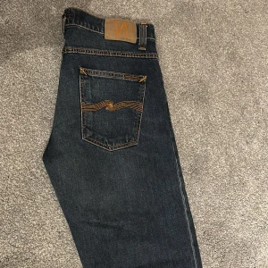 Mörkblå jeans från Nudie Jeans, W31 L32 - Snygga mörkblå jeans från Nudie Jeans med klassisk orange söm och ikonisk bakficka. Modellen har en rak passform och normal midja. Aldrig använda utom när jag prövade dem för att se om dem passade vilket dem inte gjorde. Perfekta för en stilren och avslappnad look.