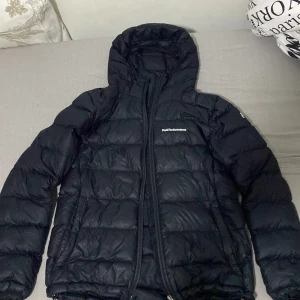 Svart dunjacka Peak Performance S - Svart dunjacka från Peak Performance med huva och dragkedja. Jackan är quiltad och har logga på bröstet samt ärmen. Perfekt för kalla dagar och har en clean, sportig look. Snygg passform och lätt att matcha med allt.och har inte använt den länge
