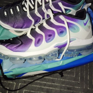 Nike Air Vapormax Plus lila/grön - Nike Air Vapormax Plus sneakers med en snygg gradient i lila, svart och mintgrön. Ovandel i syntet med vita detaljer och snörning. Genomskinlig Vapormax-sula ger futuristisk vibe och maxad komfort. Perfekta för dig som vill sticka ut med coola färger och unik design.