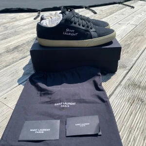 Svarta Saint Laurent sneakers - Svarta sneakers från Saint Laurent med broderad logga på sidan. Skorna har en beige platt sula, rund tå och ovandel i canvas med detaljer i skinn. Klassisk snörning och stilren design som passar till det mesta. Kommer med originalkartong och dustbag.