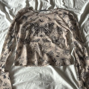 Beige mesh topp med mystiskt print - Säljer en beige mesh topp från Vanilla Cream med svart mystiskt print över hela, bland annat fjärilar, månar, rosor och kristaller. Tunn och genomskinlig med lång ärm och rund hals. Perfekt för dig som gillar unika detaljer och magisk vibe.