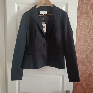 Svart kavaj från Vila, storlek L - Stilren svart kavaj från Vila i storlek L. Klassisk modell med en knapp framtill, smal krage och diskreta fickor. Perfekt för en clean och snygg look. Materialet är slätt och känns bekvämt mot huden. En tidlös bas i garderoben.