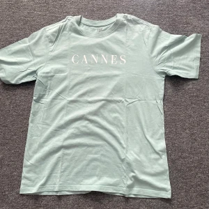 Mintgrön t-shirt från 157 med tryck - Snygg mintgrön t-shirt från 157 i 100% bomull med vit text 'CANNES' på bröstet. Klassisk passform med rund hals och korta ärmar. Perfekt för dig som gillar stilrena och fräscha plagg med enkel design.