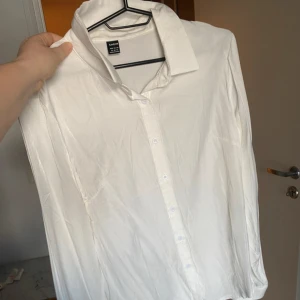 Vit skjorta från SHEIN i XL - Klassisk vit skjorta från SHEIN i storlek XL. Skjortan har lång ärm, krage och knäppning framtill med vita knappar. Materialet är lätt, så mjuk och skön och har en lite glansig finish, perfekt för en clean och stilren look.
