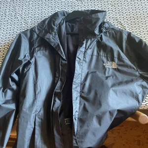 Svart vindjacka från The North Face - Snygg svart vindjacka från The North Face med huva och dragkedja framtill. Storlek 3xl men sitter som L. Endast använd ett fåtal gånger pga för stor
