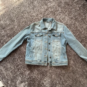 Ljusblå jeansjacka med klassisk look - Säljer en ljusblå jeansjacka med klassisk krage, bröstfickor med knappar och raka sömmar. Jackan har långa ärmar och knäppning framtill. Perfekt för dig som vill ha en tidlös och chill stil.